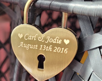 May include: A heart-shaped brass padlock featuring an engraved design with the names Carl and Jodie, two small heart icons, and the date August 13th 2016. The padlock has a central keyhole and a smooth, metallic finish with a gold-toned surface.