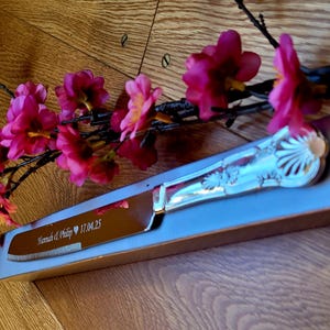 May include: A silver cake knife with an ornate handle and a serrated blade. The knife is engraved with the text "Hannah & Philip 17.04.25" and is displayed in a gray box.