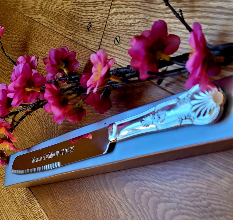 May include: A silver cake knife with an ornate handle and a serrated blade. The knife is engraved with the text "Hannah & Philip 17.04.25" and is displayed in a gray box.