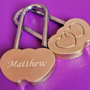 May include: Two silver heart-shaped padlocks. One padlock has the name "Matthew" engraved on it. The other padlock has two interlocking hearts engraved on it.