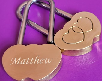 May include: Two silver heart-shaped padlocks. One padlock has the name "Matthew" engraved on it. The other padlock has two interlocking hearts engraved on it.