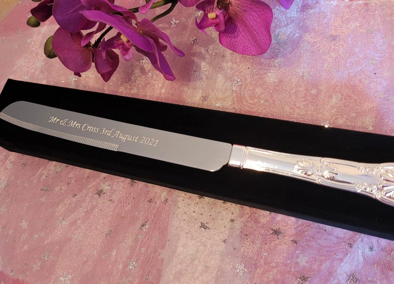 May include: Silver cake knife with a black velvet box. The knife is engraved with "Mr & Mrs Cross 3rd August 2021".