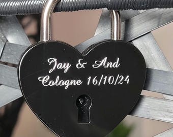 May include: A black heart-shaped padlock with the engraved text "Jay & And Cologne 16/10/24". The padlock is secured to a gray metal structure. The lock has a silver-colored shackle.