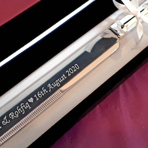 May include: A silver cake knife with a black velvet box and a white ribbon. The knife is engraved with the text "Misha & I Rohfiq &hearts; 16th August 2020".