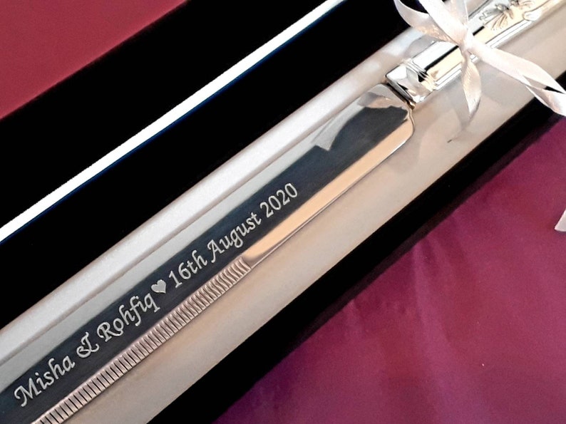 May include: A silver cake knife with a black velvet box and a white ribbon. The knife is engraved with the text "Misha & I Rohfiq &hearts; 16th August 2020".