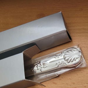 May include: A silver-plated knife with an ornate handle is displayed in a clear plastic bag inside a silver box. The box is closed and has a silver lid.