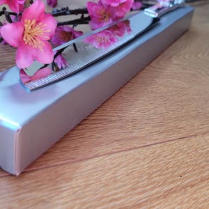 May include: A silver cake knife with a serrated edge is displayed in a silver box. The knife is resting on top of the box, with the handle extending out of the box. The box is sitting on a wooden surface.
