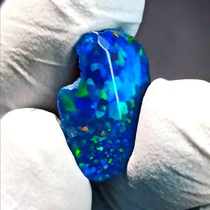 May include: A polished, irregular-shaped blue opal gemstone featuring vibrant flashes of green and small specks of orange color throughout its surface. The stone displays a deep blue base color with a high-gloss finish and a smooth, rounded edge.