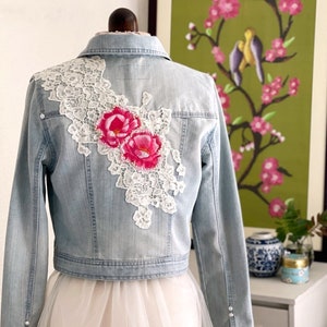 Può includere: Una giacca di jeans blu chiaro con pizzo bianco e ricamo floreale rosa. La giacca ha uno stile crop e viene indossata sopra una gonna di tulle bianca.