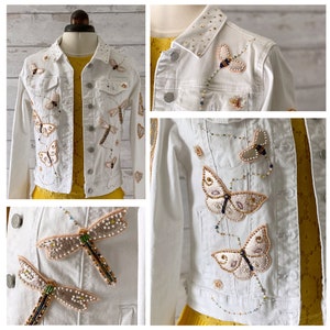 May include: A white denim jacket featuring embroidered butterflies and dragonflies. The jacket has a beaded trim and gold accents.