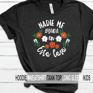 May include: Dark grey t-shirt featuring the Spanish phrase "NADIE ME AYUDA en Esta Casa" in white script, encircled by a floral design with red, white, and green flowers. The top is a crew neck style.