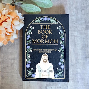 Może przedstawiać: Czarna książka zatytułowana "The Book of Mormon" ze złotymi literami i kwiatowymi akcentami. Okładka zawiera ilustrację Jezusa Chrystusa i tekst "Look Unto Christ". Ozdobne kwiaty w tle.