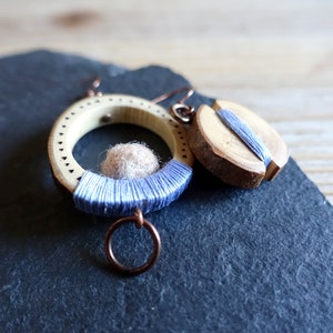 May include: A pair of wooden earrings with a circular design. The earrings are made of natural wood and feature a small, round, light brown felt ball in the center of the circle. The earrings are decorated with a blue thread wrapped around the wood.