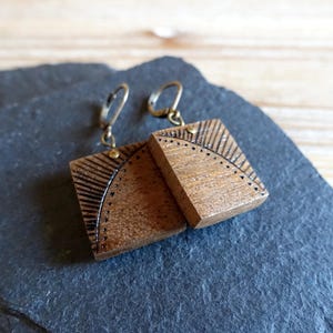 Puede incluir: Un par de pendientes de madera con un diseño geométrico. Los pendientes son de forma cuadrada y tienen un acabado de madera marrón. Los pendientes están unidos a ganchos de color dorado.
