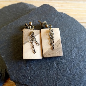 Puede incluir: Un par de pendientes de madera con un patrón de madera marrón claro y marrón oscuro. Cada pendiente tiene una pequeña cadena con un dije decorativo colgando.