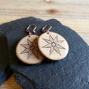 Pode incluir: Um par de brincos circulares de madeira com um design de estrela de oito pontas gravado a fogo. Os brincos são feitos de fatias de madeira natural com bordas de casca visíveis e presos a ganchos de metal na cor cobre com argolas.