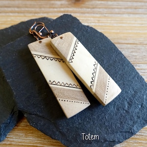 Puede incluir: Pendientes rectangulares de madera con ganchos color cobre. Los pendientes son de color madera clara con diseños geométricos en negro y marrón. La palabra "Totem" está escrita en blanco en la parte inferior.