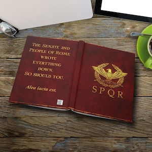May include: An open book with a burgundy cover featuring gold lettering and a gold emblem. The text reads "THE SENATE AND PEOPLE OF ROME WROTE EVERYTHING DOWN. SO SHOULD YOU. Alea iacta est. SPQR."