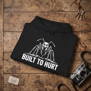 Op de afbeelding: Een zwarte hoodie met een witte grafische illustratie van een mier boven de tekst BUILT TO HURT in een vet, schreefloos lettertype. De trui is voorzien van een capuchon met trekkoord en lange mouwen in een platte lay-out.
