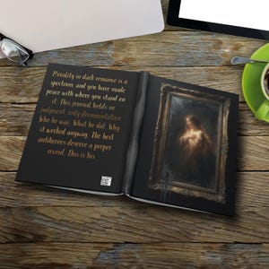 May include: An open black journal with the text "Morality in dark romance is a spectrum..." on the left page. The right page features a framed painting of a woman. The journal is on a wooden surface.