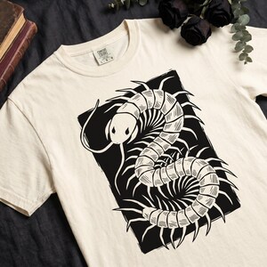 Op de afbeelding: Crèmekleurig T-shirt met een zwart duizendpootontwerp in een zwart rechthoekig kader. De duizendpoot is gedetailleerd met een gesegmenteerd lichaam en stekelige poten. Het shirt heeft een ronde hals en korte mouwen.