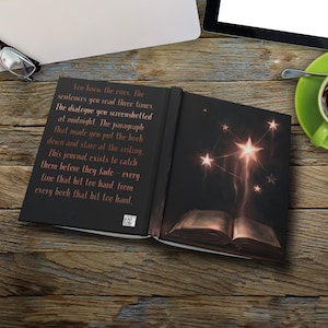 May include: Open journal with a black cover and a starry design on the right page. The left page has text in a copper color. The journal is open on a wooden surface with a cup of coffee and a tablet in the background.