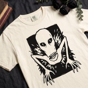 Peut inclure: T-shirt crème avec un motif noir et blanc d'une créature extraterrestre aux grands yeux et à un large sourire. Le dessin est encadré dans un rectangle noir. Le t-shirt a un col rond et des manches courtes.