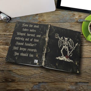 May include: Open book with a dark, textured cover. The left page has gothic text: "Even the devil takes notes." The right page features a skeleton with wings, horns, and a spear. The book's theme is dark and gothic.