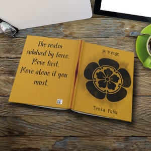 Peut inclure: Un livre ouvert avec une couverture et des pages jaune moutarde. La page de gauche contient le texte : « Le royaume soumis par la force. Agissez en premier. Agissez seul si vous devez. » La page de droite a un motif floral noir et du texte japonais.