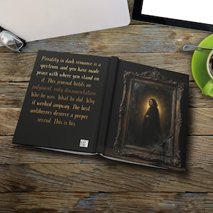 May include: Open journal with black cover featuring text about dark romance and a framed portrait. The portrait depicts a figure in a dark cloak. The journal is open on a wooden surface.