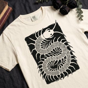 Op de afbeelding: Crèmekleurig T-shirt met een groot zwart-wit duizendpootontwerp. De afbeelding is een gestileerde illustratie van het insect, met ingewikkelde details. Het shirt heeft een klassieke ronde hals en korte mouwen.