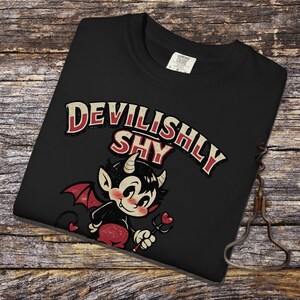 Puede incluir: Camiseta negra con el texto "DEVILISHLY SHY" en rojo y blanco y una ilustración de diablo de dibujos animados. El diablo tiene alas rojas y una cola, sosteniendo un tridente en forma de corazón. La camiseta está doblada sobre una superficie de madera.
