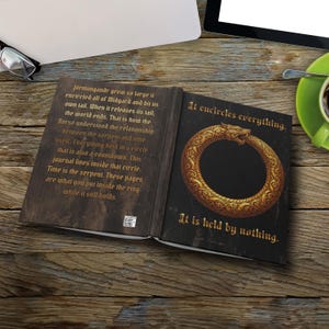 May include: An open journal with a dark cover featuring a gold Ouroboros serpent graphic on the right page and text on the left page. The text reads: Jörmungandr grew so large it encircled all of Midgard and bit its own tail. It encircles everything.