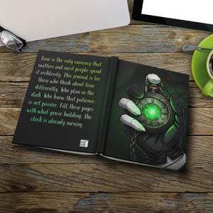 May include: Open journal with a green and black design. The right page features a detailed illustration of a gloved hand holding a glowing green pocket watch. The left page has white text on a black background. The journal is on a wooden surface.