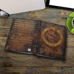 Könnte beinhalten: Ein aufgeschlagenes Notizbuch mit dunkelbraunem, strukturiertem Einband und einem goldenen Ouroboros-Schlangenmotiv. Die linke Seite enthält gedruckten Text über Jörmungandr, während auf der rechten Seite It encircles everything. It is held by nothing. steht.