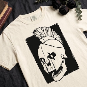 Könnte beinhalten: Cremefarbenes T-Shirt mit einer schwarz-weißen Grafik eines Schädels mit Helm und Irokesenschnitt. Der Schädel ist vor einem schwarzen rechteckigen Hintergrund platziert. Das Shirt hat einen Rundhalsausschnitt und kurze Ärmel.