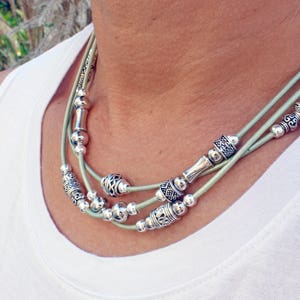 Puede incluir: Un collar de varias capas con cuentas plateadas y elementos decorativos ensartados en tres hebras de cordón verde claro. El collar se usa sobre una camiseta blanca, con cuentas de diferentes tamaños y diseños.