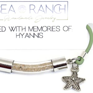 May include: A handmade bracelet with a clear tube filled with sand, silver-tone metal accents, and a starfish charm. The bracelet has a green cord and the text "FILLED WITH MEMORIES OF HYANNIS".