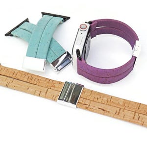 May include: An assortment of Apple Watch bands in various colors and materials. The bands include a light teal cork band, a natural cork band, and a purple band. Each band has a silver clasp and black connectors.