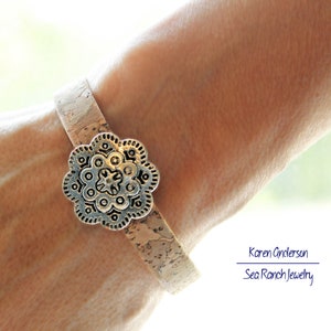 May include: A cork bracelet with a silver-colored, flower-shaped clasp. The bracelet is light brown and appears to be made of cork. The clasp has an intricate design. The bracelet is worn on a wrist.