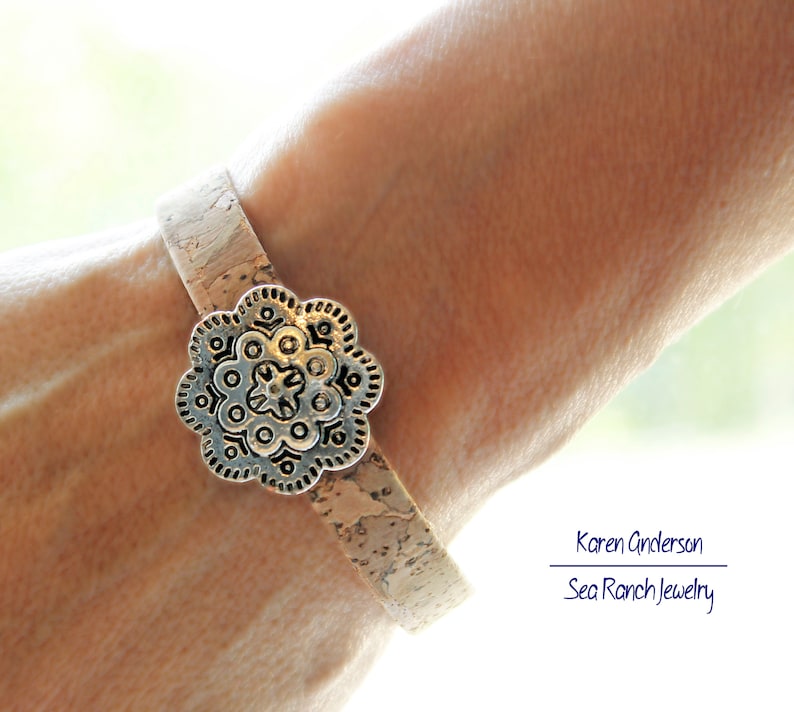 May include: A cork bracelet with a silver-colored, flower-shaped clasp. The bracelet is light brown and appears to be made of cork. The clasp has an intricate design. The bracelet is worn on a wrist.