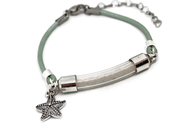Pode incluir: Uma pulseira com uma faixa de couro verde claro e um tubo transparente cheio de areia. A pulseira tem detalhes em metal prateado e um pingente de estrela do mar. A corrente ajust&aacute;vel tamb&eacute;m &eacute; prateada.