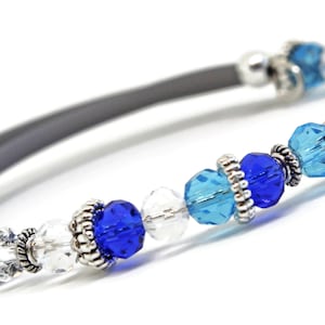 May include: A beaded bracelet featuring a gray band and an assortment of blue and clear crystal beads. The bracelet has silver-colored metal accents. The beads are various shades of blue, including light blue and deep blue.