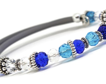 May include: A beaded bracelet featuring a gray band and an assortment of blue and clear crystal beads. The bracelet has silver-colored metal accents. The beads are various shades of blue, including light blue and deep blue.