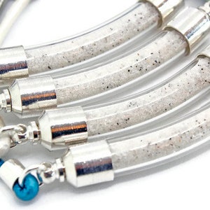 Pode incluir: Close-up de v&aacute;rias pulseiras. Cada uma tem um tubo de vidro transparente e curvo preenchido com areia branca e detalhes em metal prateado. As pulseiras t&ecirc;m cord&otilde;es de couro coloridos em tons de azul, verde e castanho.