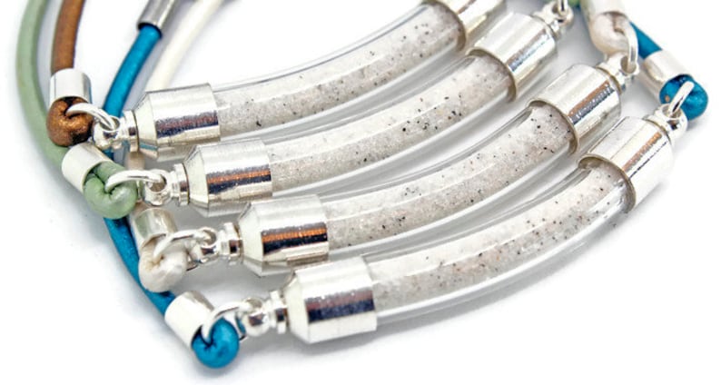 Pode incluir: Close-up de v&aacute;rias pulseiras. Cada uma tem um tubo de vidro transparente e curvo preenchido com areia branca e detalhes em metal prateado. As pulseiras t&ecirc;m cord&otilde;es de couro coloridos em tons de azul, verde e castanho.