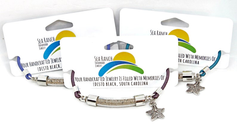 Pode incluir: Tr&ecirc;s pulseiras com um tubo transparente cheio de areia, conectadas por metal prateado. Cada pulseira tem um cord&atilde;o de cor diferente: azul, roxo e azul-petr&oacute;leo. Um berloque de estrela do mar pende de cada pulseira. A embalagem diz "Sea Ranch Handmade Jewelry".