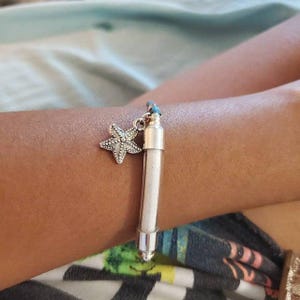Pode incluir: Uma pulseira de prata com um berloque de estrela do mar e um frasco transparente preenchido com uma subst&acirc;ncia branca. O berloque de estrela do mar &eacute; detalhado com pequenos enfeites. A pulseira &eacute; usada em um bra&ccedil;o.