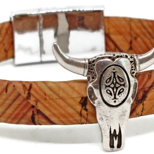 May include: A cork bracelet with a silver-colored bull skull charm. The bracelet band is made of natural cork material with a textured pattern. The bull skull charm has detailed features and a magnetic clasp.