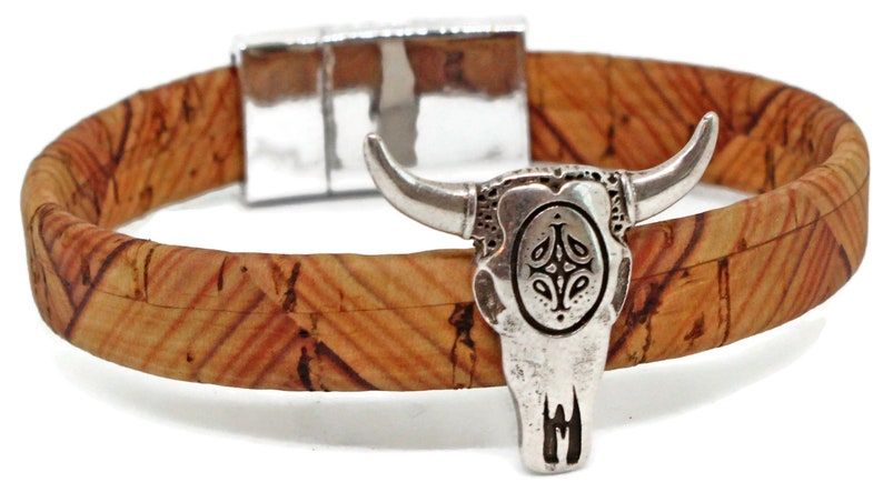 May include: A cork bracelet with a silver-colored bull skull charm. The bracelet band is made of natural cork material with a textured pattern. The bull skull charm has detailed features and a magnetic clasp.